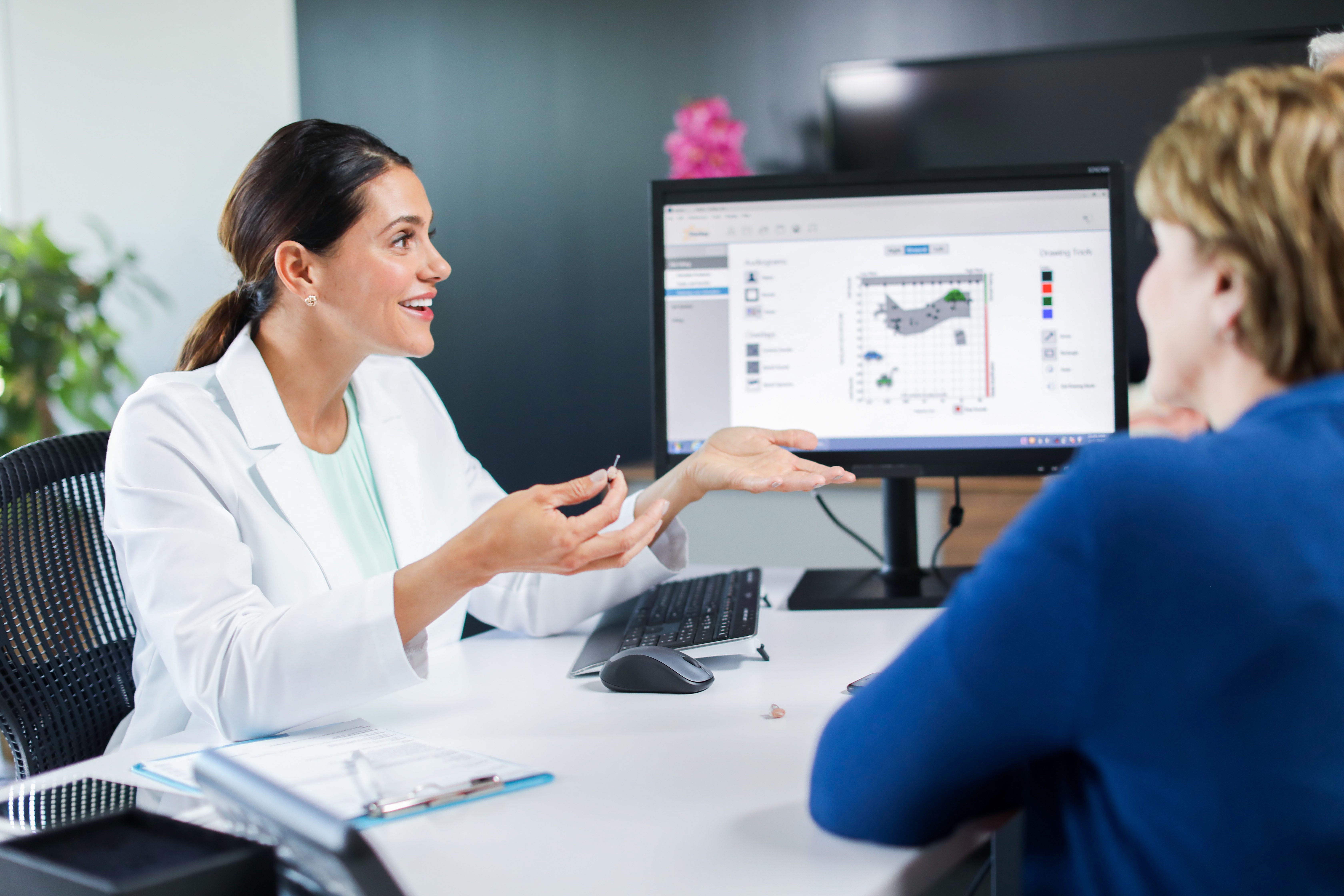 Woman in lab coat holding a hearing aid talking to a patient